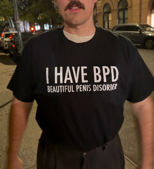 A man with a mustache stands on a city street at night wearing a black t-shirt that reads 'I HAVE BPD BEAUTIFUL PENIS DISORDER'.