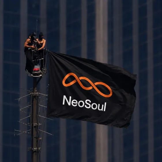 Two people in dark clothing are perched atop a tall pole or antenna against a blue skyscraper, unfurling a large black flag with an orange infinity symbol and the word 'NeoSoul'.