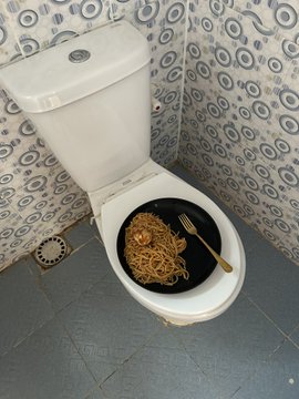 The image shows a small white toilet with a plate of spaghetti sitting inside it, which is surrounded by various bathroom fixtures like a sink and a shower head. The background features a wall with blue and silver geometric patterns that add an interesting visual element to the scene. A caption next to the photo reads: "don't ask me how, but it's cause if her BTC is at $69k". This statement implies that someone has posted this image for attention or amusement rather than actual purpose or context.