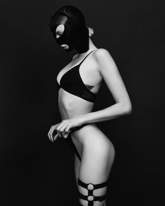 A black-and-white photo shows a figure in profile wearing a black Catwoman-style mask, black bikini, and thigh harness with garter straps against a dark background. The edgy, mysterious aesthetic pairs with the "Gm guys🖤🤘🏻" greeting as a stylish good morning post.