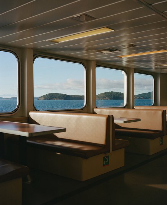 The interior of a ferry or boat features empty booth seating with brown cushions and tables, looking out through large windows at a calm sea and forested islands.