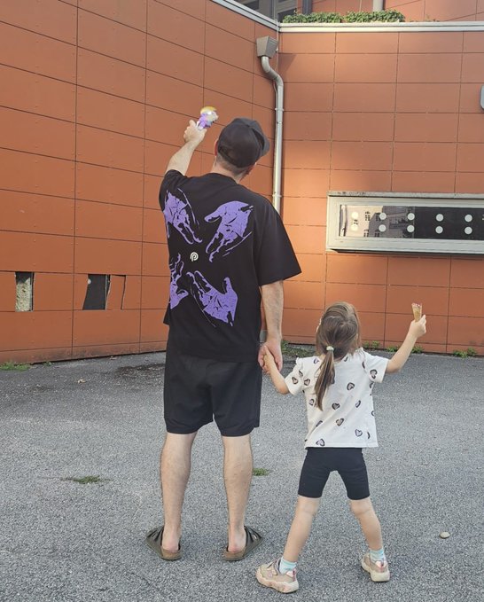 A man and a young girl holding hands outdoors against an orange tiled wall, with the man wearing a black t-shirt with purple graphics and holding up a small toy, while the girl wears a white shirt with heart patterns.