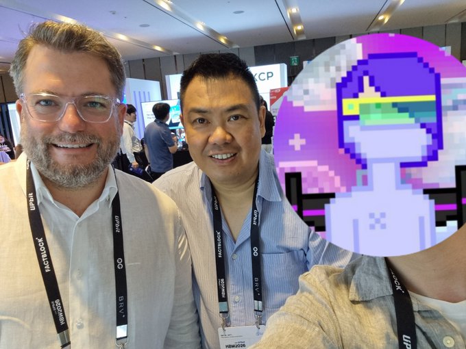 Two men smiling at a conference, one wearing a 'HASHLOCK' badge reading 'Hywit Energy', with a pixel art avatar covering the third person's face on the right.