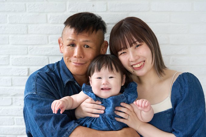 A smiling Japanese family of three—father, mother, and baby girl—pose together in matching denim outfits against a white brick wall, celebrating their daughter's 1st birthday alongside her father's birthday on July 4th.