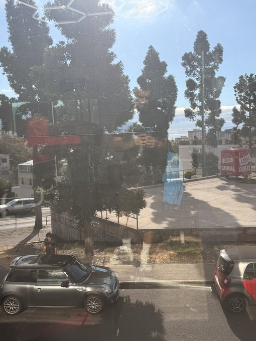 The image, taken through a reflective window, shows a person standing next to a dark Mini Cooper and a red car parked on a street, with a delivery truck labeled "CORAL" visible across the way. The reflective glass surface creates a ghostly overlay of the photographer and surroundings, adding a mysterious, transparent quality that visually echoes the anonymity and trust considerations of sharing a cryptocurrency wallet address publicly.