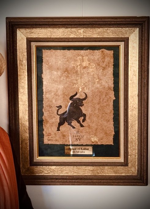 A framed art piece titled "699478" (the Bitcoin Law SV) featuring a black bull silhouette on parchment-like paper, displayed with a plaque reading "House of Rallos El Salvador" in honor of El Salvador's historic Bitcoin Law adoption.