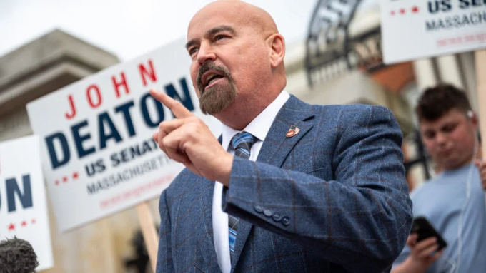 A man in a blue plaid suit gestures with his finger raised while speaking in front of campaign signs for 'John Deaton US Senate Massachusetts'.