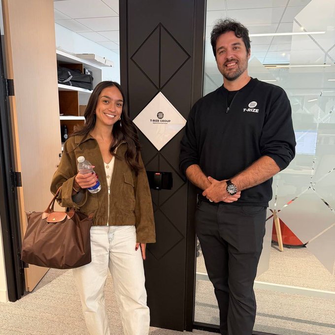 Carina poses alongside JL Pellerin at the T-Rize Group office during a visit to discuss collaboration on growing the Canton Network ecosystem. The image captures the two standing by a door labeled "T-RIZE GROUP," reflecting the productive partnership between their teams.