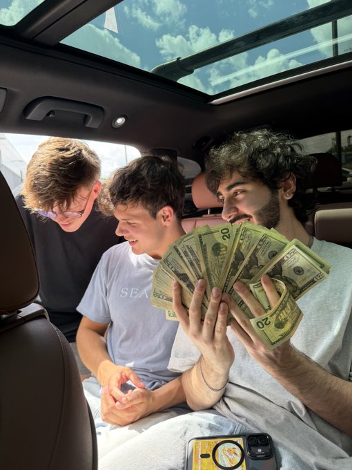 Three young men sit inside a car with a panoramic sunroof, as the man on the right excitedly fans out a large stack of $20 bills while the others lean in to look. The scene captures a celebratory cash giveaway moment tied to the "$ANSEM" post, with a phone in the foreground ready to record the distribution.