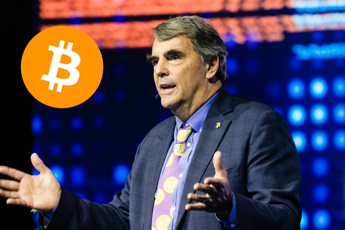 Billionaire Tim Draper is shown mid-speech at a conference, gesturing with his hands while wearing a navy suit, blue shirt, and a distinctive yellow Bitcoin-themed tie. A large Bitcoin logo is overlaid on the left side of the image, visually reinforcing his bold prediction about Bitcoin eventually replacing traditional currency and triggering a fundamental shift in the global financial system.