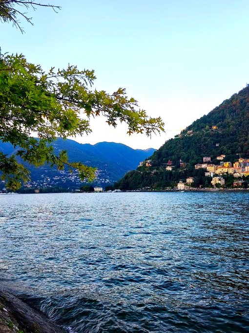 A scenic view of an Italian lake with rippling blue water, mountains in the background, and a hillside town with colorful buildings on the right, framed by a tree branch with green leaves.