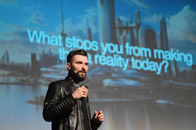 A bearded man in a black leather jacket speaks into a microphone on stage with the text 'What stops you from making ___ a reality today?' displayed behind him against a futuristic city backdrop.