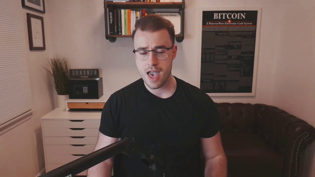 The man with glasses stands holding a microphone next to a couch, appearing to be singing or speaking into the microphone during a live streaming session. There are also several books visible in the background, suggesting that he might have some knowledge about cryptocurrencies like Bitcoin or other financial topics. The image appears to be promoting Bitcoin and related themes through various elements such as singing, a cozy setting with a couch, and books, indicating an attempt at a podcast or video showcasing more information on Bitcoin and its history.