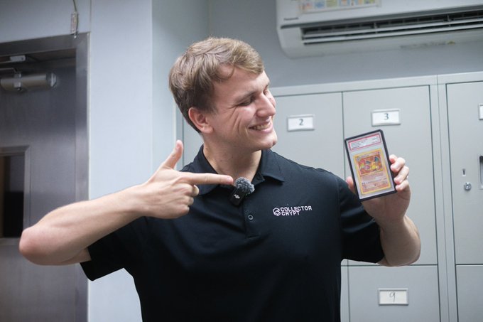 A smiling man in a black 'Collector Crypt' polo shirt points to a Pokémon trading card he's holding in a room with grey lockers.