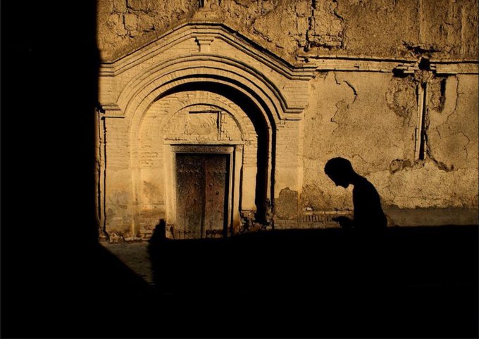 A silhouette of a person reading is cast in shadow against a warmly lit, weathered stone wall featuring an arched doorway and a mounted cross.