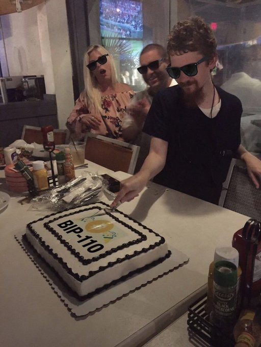 A group of people wearing sunglasses celebrate at a table with a birthday cake decorated with 'BIP-110' and a Bitcoin logo.