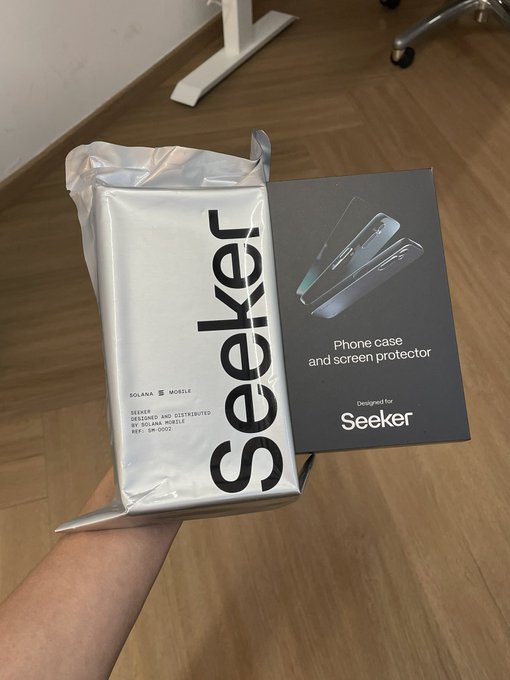 A hand holding a silver Seeker-branded package alongside a dark box labeled 'Phone case and screen protector - Designed for Seeker' over a wooden floor.