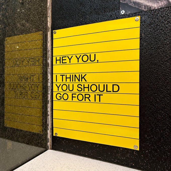 A bright yellow sign mounted on a black wall displays the encouraging message "HEY YOU, I THINK YOU SHOULD GO FOR IT," which aligns perfectly with the post's theme of challenging yourself every day, serving as a bold visual reminder to embrace ambition and take action.