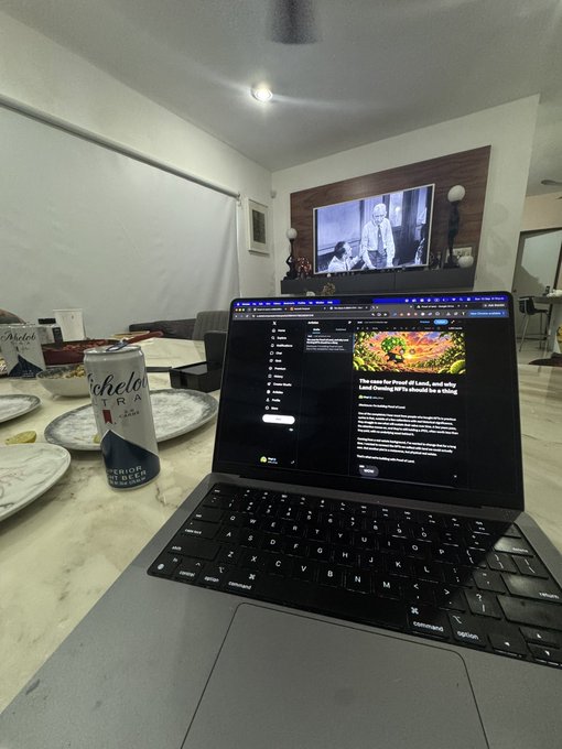 An open laptop on a table displays a webpage featuring an aerial landscape photo and the headline "The coast the flood of Lixuri, and why I tried turning 60% to almonds for a living", with a can of Michelob Ultra and a TV showing a scene in the background.