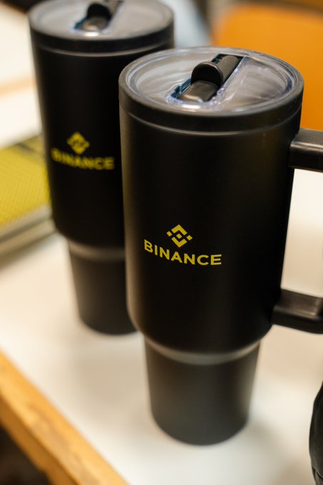 Two matte black tumblers with clear lids sit on a light surface, both prominently branded with the yellow Binance logo and name. The post's playful "downloading morning energy" caption fits perfectly—these sleek crypto-branded travel mugs look like the kind of essential gear you'd grab to fuel up before a busy day in the markets.
