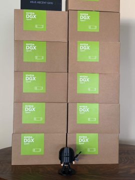 A stack of NVIDIA DGX boxes (appearing to be ten boxes arranged in two columns) with green labels, with a small figurine placed at the bottom for scale.
