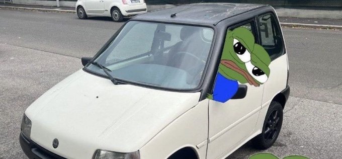 A small white car is parked on a street with a sad Pepe the Frog cartoon character edited into the driver's side window.