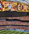 A large stadium display showing the word 'GOLD' in a yellow and red graphic, overlooking a baseball field and crowded stands.