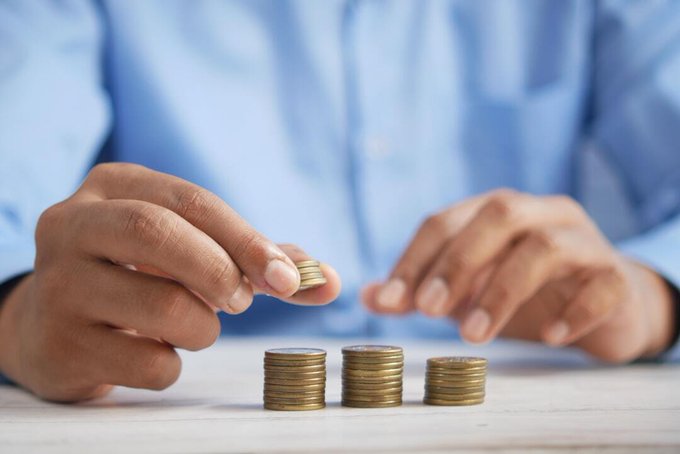 A person in a blue shirt stacks and counts gold-colored coins, arranging them into three separate piles on a light wooden surface.