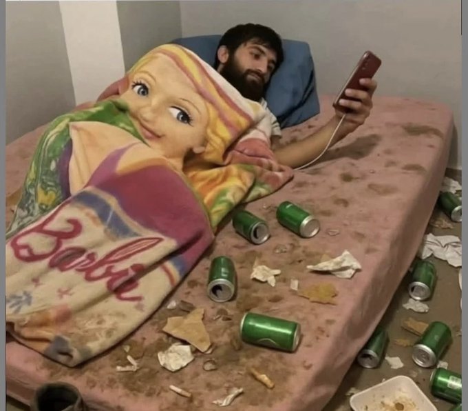 A bearded man lies on a dirty, stained mattress in a messy room, wrapped in a Barbie blanket and looking at his phone, surrounded by empty green beer cans and trash.