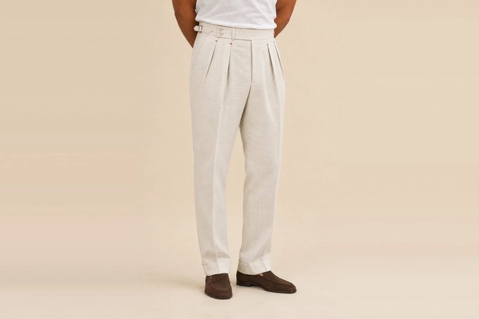 A product photo of a man wearing cream-colored high-waisted pleated trousers paired with brown suede loafers and a white shirt against a beige background.