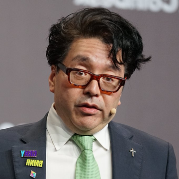 A man with dark hair and red-framed glasses wearing a dark suit, white shirt, and green tie, with a "WE THE AMMO" pin on his lapel and a headset microphone, appears to be speaking at a conference.