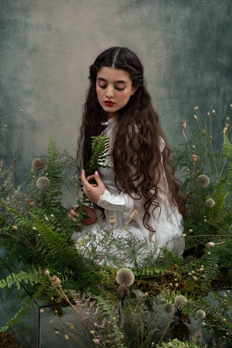 A portrait of a woman with long wavy brown hair in a white Victorian-style dress, sitting among ferns and flowers with her eyes closed.