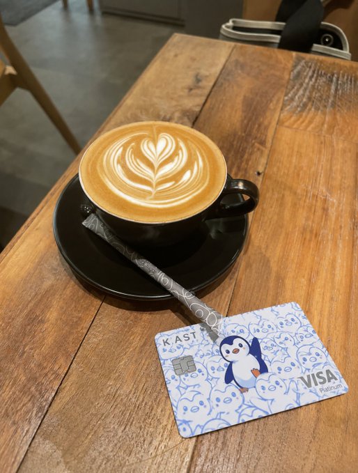 The image features a wooden table with various objects placed on top, including a cup of coffee, a payment card, and a penguin sticker. The coffee appears to be latte art style, adding an artistic touch to the scene. A payment card is also present on the table, suggesting that this could be a casual setting where someone might enjoy their coffee while making a purchase using the Visa card. The post seems to be promoting the idea of integrating modern finance into everyday life and experiences, such as enjoying a cup of coffee with a payment card in hand.