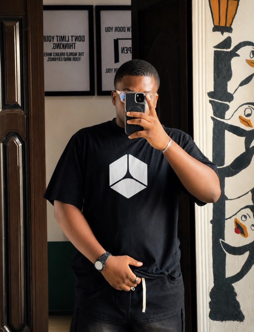 A mirror selfie of a man wearing a black t-shirt featuring a geometric cube logo, with framed motivational text visible in the background.