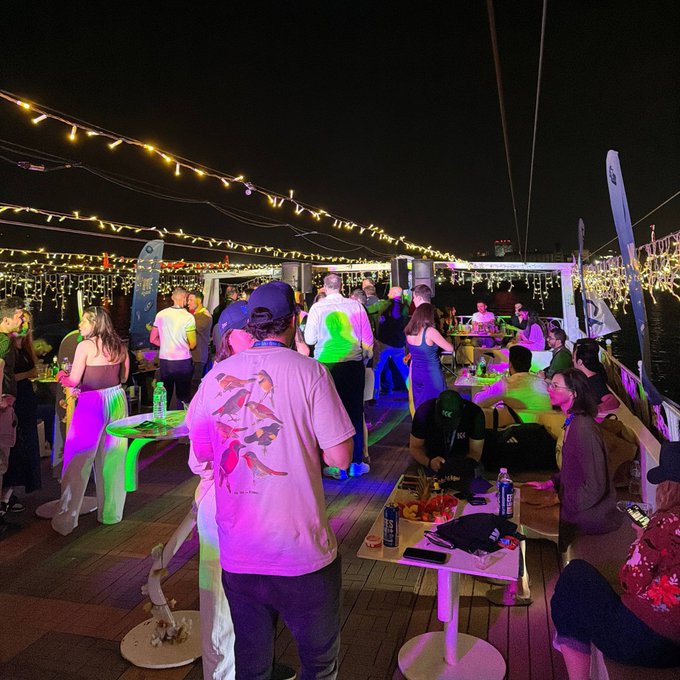 The image shows a lively scene at a party, with people standing around tables filled with food and drinks. The guests are enjoying themselves as they mingle and socialize under the lights of colorful string lights hanging above them. A boat can be seen in the background, adding to the festive atmosphere. The post about the Sui Yacht party suggests that this gathering is taking place on a boat or at an event with a boating theme.