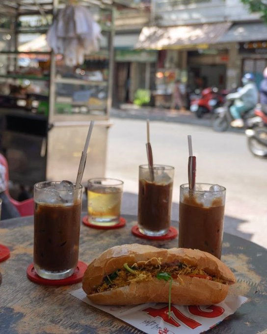 A street-side café scene shows a Vietnamese bánh mì sandwich on a plate with multiple glasses of iced coffee arranged on a wooden table, with motorbikes and a shop visible in the background.