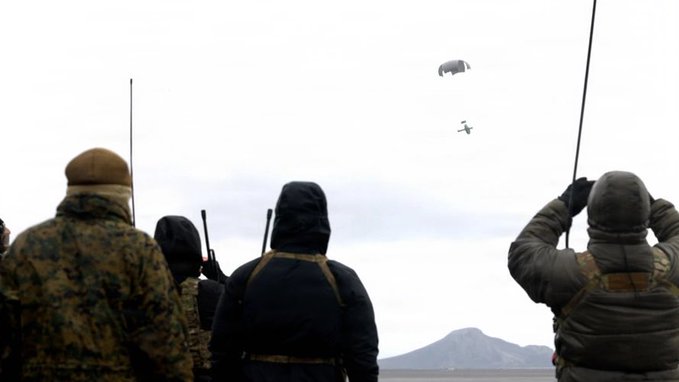 People in military gear watch as a small drone and parachute float through the sky during an aerial resupply operation. The scene captures the onlooking personnel observing the critical medical supplies being delivered by the Grasshopper drone, aligning with the trilateral exercise between the UK and Norway involving Ondas/Dzyne's technology ($ONDS).