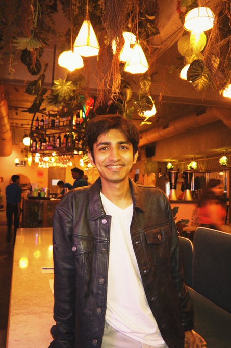 A young man wearing a black leather jacket over a white t-shirt, smiling in a warmly lit restaurant with decorative plants and hanging lamps.