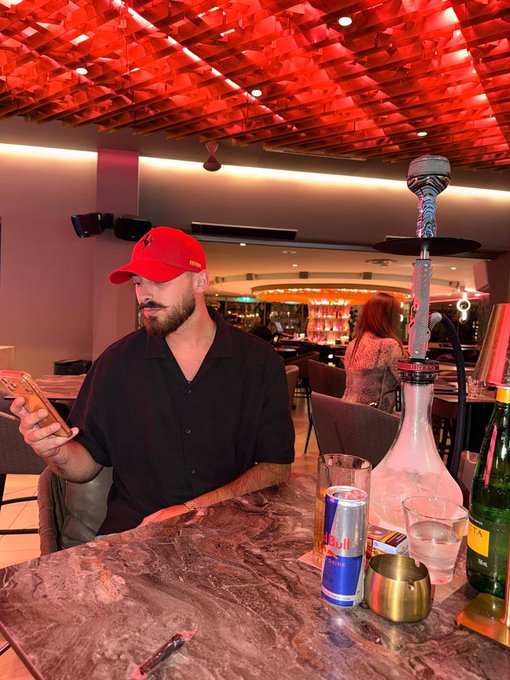 A man wearing a red hat and dark shirt sits at a bar featuring a Red Bull can and a hookah, appearing to be on his phone. The caption "We're about to see the biggest crypto bull run in human history" contrasts with the relaxed lounge atmosphere, suggesting he's perhaps ignoring the excitement in favor of personal downtime.