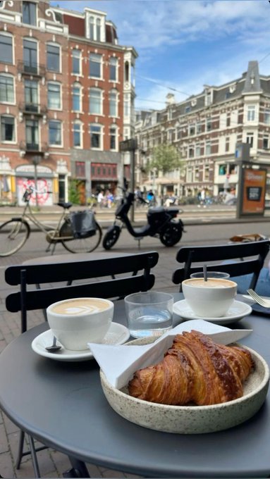 A morning coffee and croissant at a sidewalk café, with classic Amsterdam canal houses, bicycles, and a scooter visible in the background. The image perfectly captures the "GM from Amsterdam" post, showcasing a quintessential Dutch morning scene.