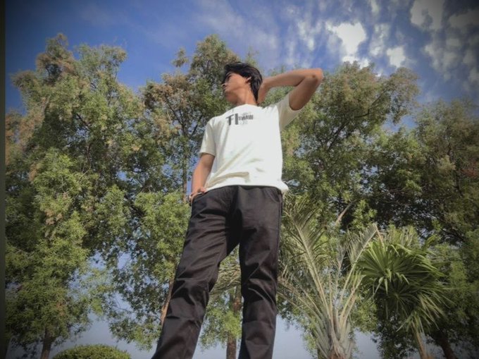 A low-angle photograph of a person wearing a white 'Thrasher' t-shirt and black pants, standing with one hand behind their head against a backdrop of trees and a cloudy blue sky.