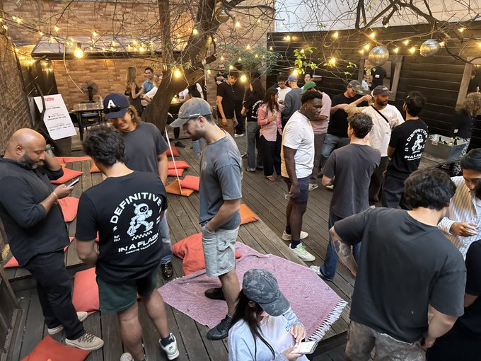 A group of builders gathers on a wooden deck adorned with string lights at the opening night of the Runtime hackathon, with attendees wearing shirts featuring "DEFINITIVELY IN A FLASH" text. Cushions and blankets are scattered across the floor as participants socialize and prepare for the upcoming September 19th submissions.