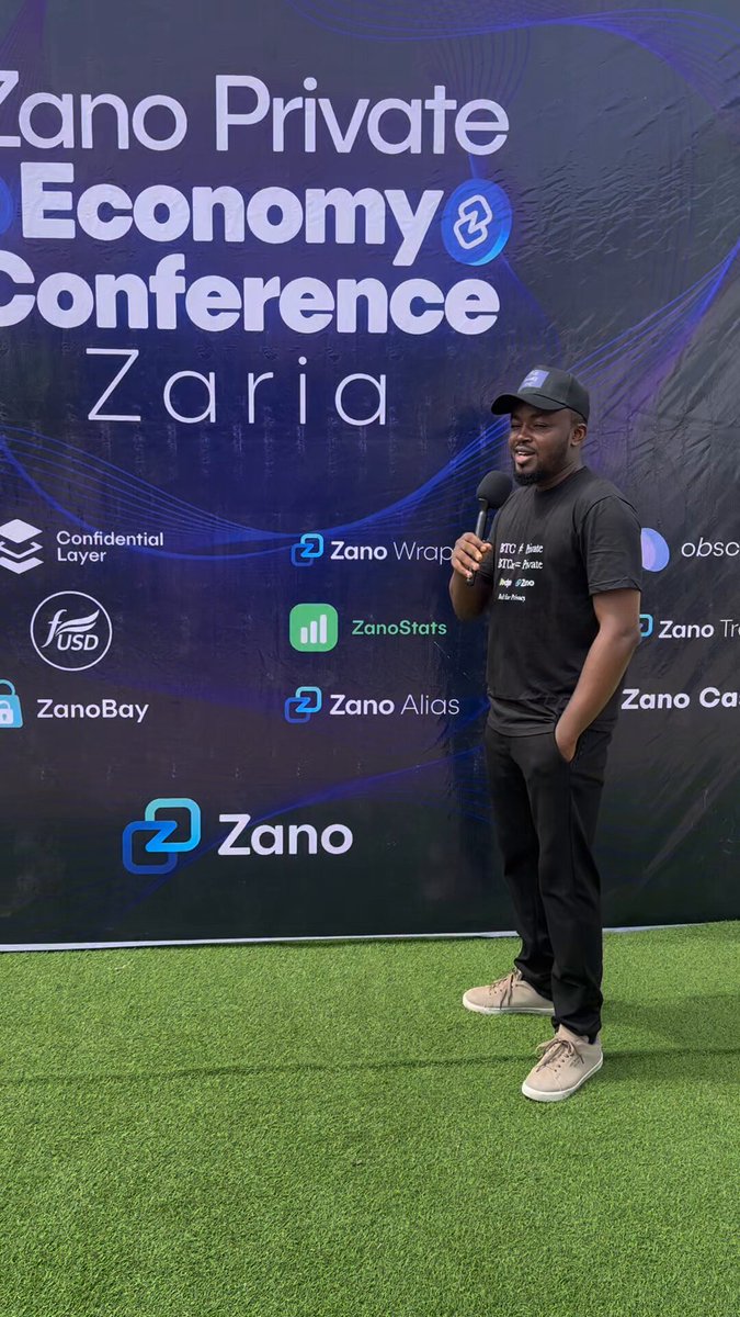 The image features a man standing next to a banner for the ZanoAfrica Private Economy Conference. The man appears to be giving a speech or presentation, as he is holding a microphone and wearing a black shirt with white text. The background of the image shows a green grass field, which contrasts with the dark blue sky above. The overall atmosphere suggests that this event is an important gathering for those interested in financial privacy and digital freedom.
