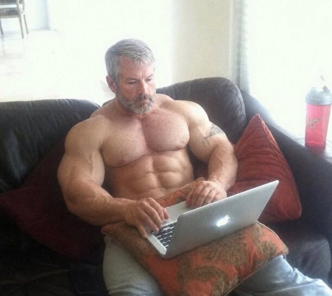 A shirtless, muscular older man with gray hair and a beard sits on a dark leather couch using a MacBook laptop resting on an orange patterned pillow, with a pink water bottle nearby. The caption "Time to lock in bros." humorously suggests he's intensely focused and ready to grind, with his serious expression and flexed physique reinforcing the motivational "locked in" mentality.
