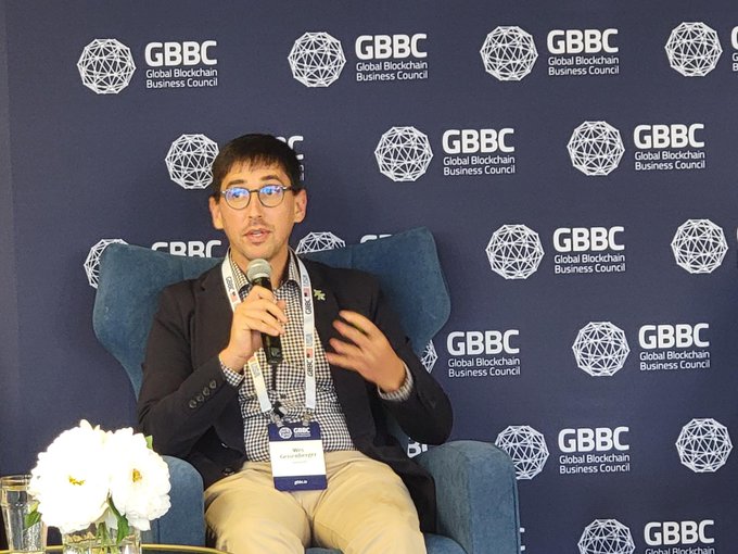 A young man wearing glasses and a dark blazer speaks into a microphone while seated on a blue chair, with a backdrop displaying the 'GBBC Global Blockchain Business Council' logo repeated across it.