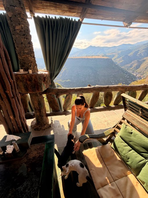 A woman sits on a rustic wooden terrace petting a black and white cat, with scenic mountains and a blue sky visible in the background.
