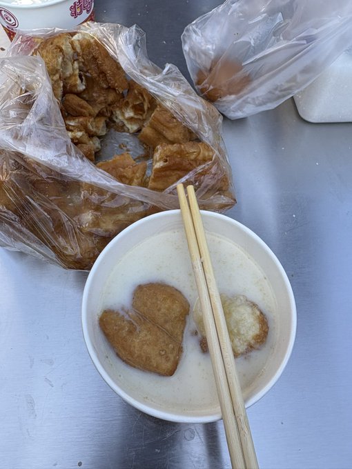 A simple meal of fried dough pieces in a bowl of milky white soup with chopsticks resting on top, alongside a plastic bag containing more of the fried dough on a metal table.