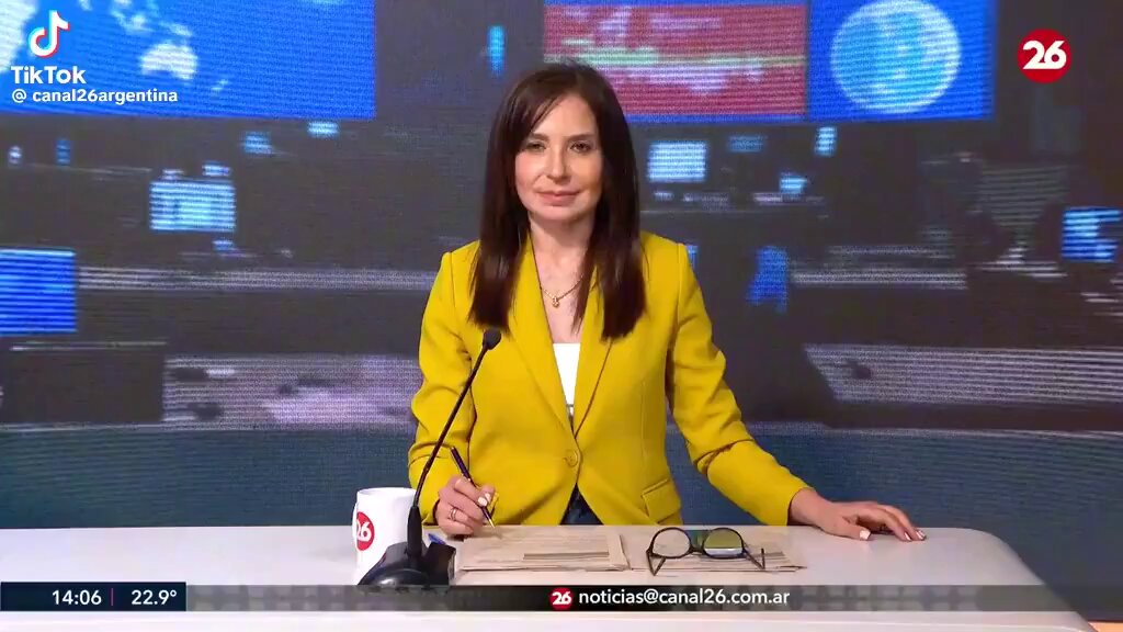 The image shows a woman wearing a yellow blazer sitting behind a desk, with a TV screen in front of her displaying information related to a political event or news story. The TV screen also features a map of the world, which could be part of the content being discussed on the show. The woman appears to be engaged and focused on the topic at hand, possibly reporting from an important event or discussing a significant issue.