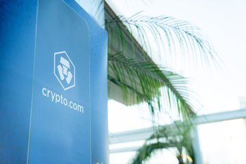 The image shows a blue sign with white text that reads "Crypto.com" against a white background. The sign is located next to a palm tree, suggesting an outdoor setting or event space. There are also some people visible in the scene, possibly attending the event or interacting with the sign.