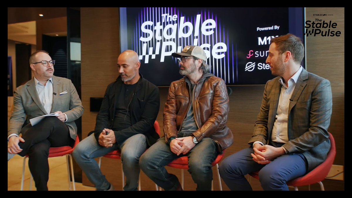The image shows four men sitting around a table with their legs crossed, engaged in conversation. They are positioned against a wall that has a large screen displaying "The Stable Pulse" behind them. The group appears to be discussing something related to stablecoins or the cryptocurrency market.